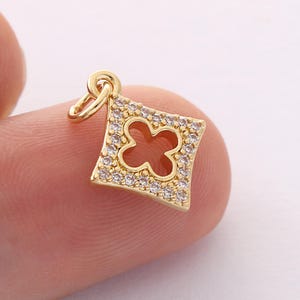 May include: A gold-colored charm with a four-leaf clover design. The charm is square-shaped with a clover cutout in the center and is encrusted with small, clear crystals around the edges. It has a small loop at the top for attachment.