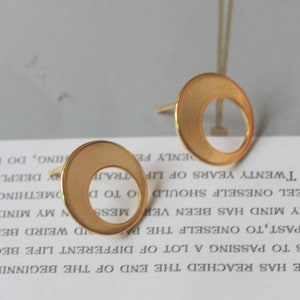 May include: A pair of gold-colored stud earrings. Each earring features a circular design with a crescent-shaped cutout. The earrings have a matte finish and are set against a blurred background.