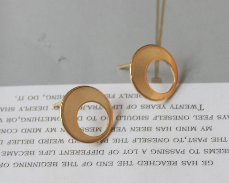 May include: A pair of gold-colored stud earrings. Each earring features a circular design with a crescent-shaped cutout. The earrings have a matte finish and are set against a blurred background.