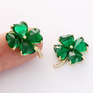 May include: Two gold-toned four-leaf clover charms with green gemstone petals. Each charm has a curved stem embellished with small, clear crystals. The clover leaves are heart-shaped and faceted, reflecting light.
