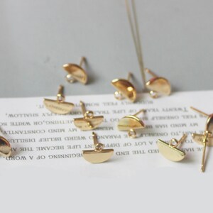 May include: A group of eight gold-toned earring findings with a half-circle shape and a small loop at the top. The findings are scattered on a white surface with text.