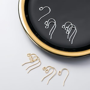 May include: A close-up of two sets of earring hooks. One set is silver and the other is gold. The hooks are displayed on a black and gold plate and a white surface.