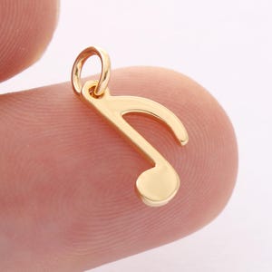 Könnte beinhalten: Ein goldfarbener Musik-Achtelnoten-Anhänger. Der Anhänger hat einen glänzenden, polierten Goldton mit einer Schlaufe oben zum Befestigen an einer Kette oder einem Armband. Die Note hat einen abgerundeten Kopf und einen gebogenen Schwanz.
