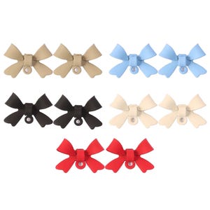 Puede incluir: Un surtido de adornos para zapatos en forma de lazo, en varios colores, incluyendo beige, azul claro, negro, crema y rojo. Cada adorno cuenta con un pequeño accesorio circular para una fácil colocación en el calzado. Los adornos están dispuestos en pares.