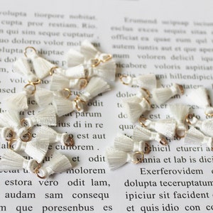 May include: A string of white tassel charms with gold jump rings. The tassels are made of soft, fluffy white yarn and are attached to the rings with a knot. The charms are strung together with a gold chain.