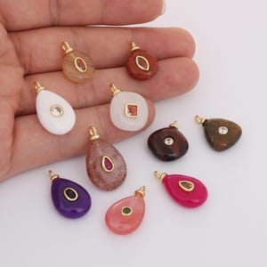 Può includere: Una collezione di ciondoli con pietre preziose in varie forme e colori, tra cui bianco, marrone, rosa e viola. Ogni ciondolo ha un'ansa color oro e presenta un accento di pietra preziosa o cristallo.