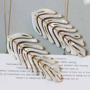May include: A pair of white, leaf-shaped earrings with gold accents. The earrings are made of a textured material and have a delicate, feminine design.