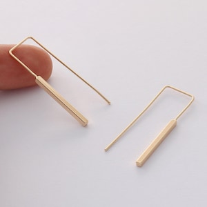 Op de afbeelding: Een paar minimalistische gouden oorbellen met een dunne rechthoekige staaf die aan een dunne draadlus hangt.