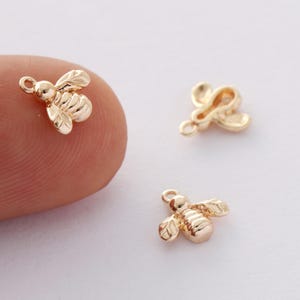 May include: Close-up of three small, gold-colored bee charms. Each charm features detailed wings and body segments, with a small loop at the top for attaching to jewelry. The bees are set against a soft, neutral background.