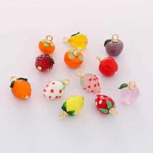 Op de afbeelding: Een verzameling kleurrijke fruitvormige bedels met goudkleurige lussen. De bedels zijn onder andere aardbeien, sinaasappels, citroenen, appels en ander fruit. De bedels zijn gemaakt van glas of hars en zijn in verschillende tinten rood, oranje, geel en roze.