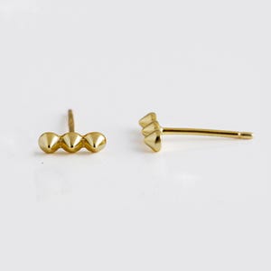 May include: A pair of gold stud earrings. One earring features three small, rounded cones in a row. The other earring features three small, rounded cones stacked vertically.
