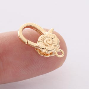 Puede incluir: Un cierre de langosta dorado con un diseño floral. El cierre está hecho de metal y presenta una flor decorativa en la parte delantera. El cierre está diseñado para asegurar piezas de joyería.