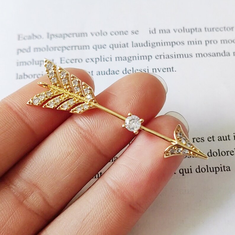 May include: A gold arrow charm with clear crystal accents. The arrow is adorned with small, sparkling crystals that create a dazzling effect.