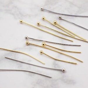 May include: Assortment of gold and silver head pins with round ball heads. The pins are scattered on a white marble surface.
