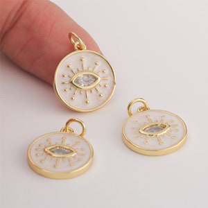 May include: Three gold-tone pendants with white enamel and a clear crystal eye design. Each pendant features a circular shape with a raised eye design and a small loop at the top for attaching to a chain or cord.