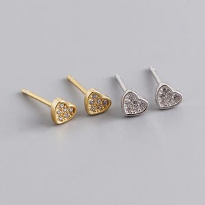 Può includere: Due paia di orecchini a lobo a forma di cuore. Un paio è dorato con pietre trasparenti, l'altro è argentato con pietre trasparenti. Gli orecchini sono esposti su uno sfondo grigio.