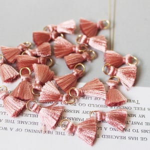 May include: A collection of small, blush-pink tassels with gold-colored loops. Each tassel has a bow-like top and a fringe of silky threads. The tassels are scattered on a light-colored surface with faint text visible.