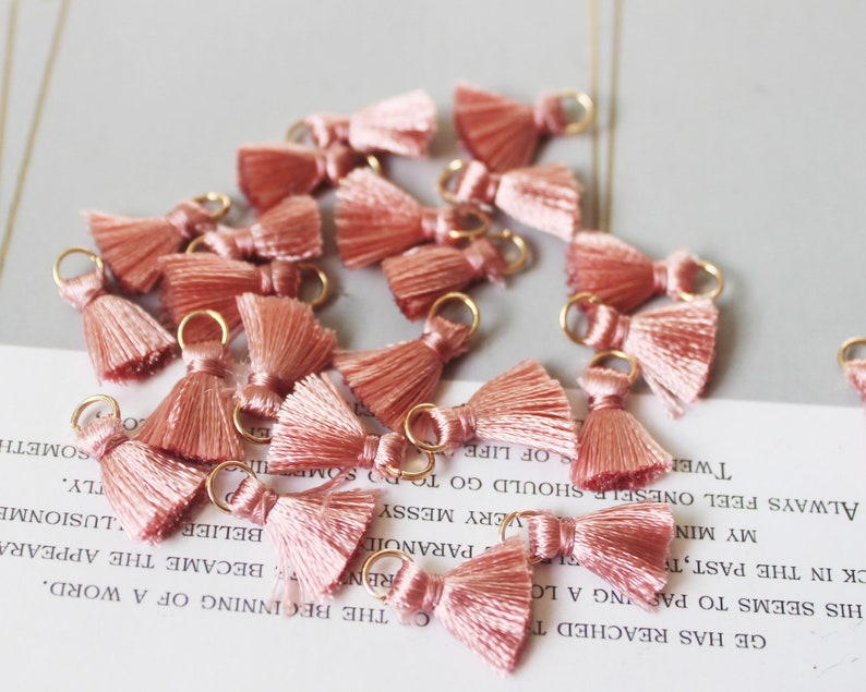 May include: A collection of small, blush-pink tassels with gold-colored loops. Each tassel has a bow-like top and a fringe of silky threads. The tassels are scattered on a light-colored surface with faint text visible.