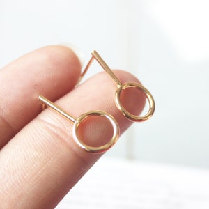 May include: A pair of gold-toned minimalist earrings with a small circle design. The earrings have a simple, modern style.