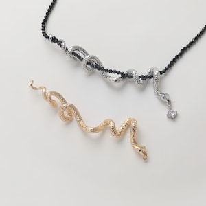 Op de afbeelding: Twee slangen-vormige kettingen, een zilveren met een zwarte kralenketting en een heldere kristallen hanger, en een gouden. Beide kettingen liggen op een witte achtergrond.