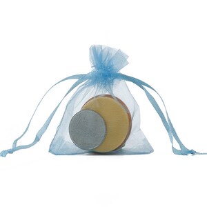 Op de afbeelding: Een lichtblauwe organza-geschenkzak met een trekkoord. De zak bevat drie ronde, platte voorwerpen: een zilverkleurige schijf, een lichtbruine schijf en een donkerbruine schijf.