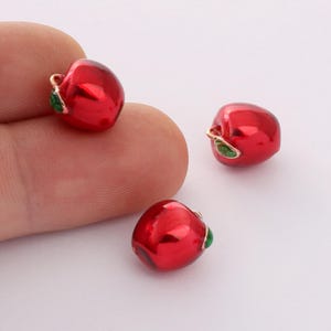 May include: Three red apple charms with green leaves. The charms are made of metal and have a shiny finish.