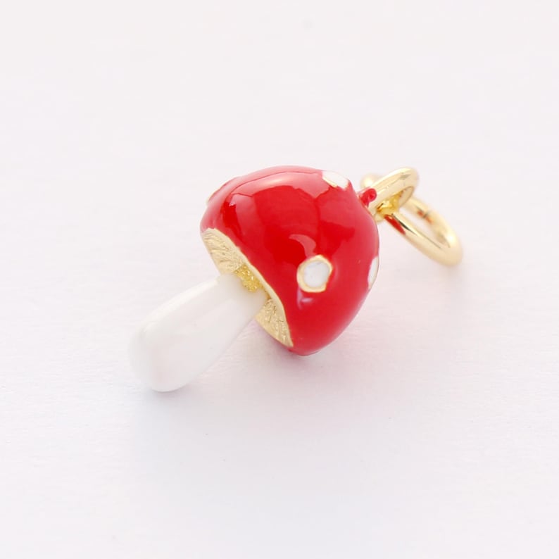 May include: A red and white enamel mushroom charm with a gold-tone finish. The charm has a white stem and a red cap with white polka dots.