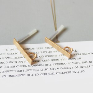 May include: Two gold-colored bar earrings with a small loop at the end. The earrings have a post for insertion into the ear. The background is a light gray surface with text from a book.