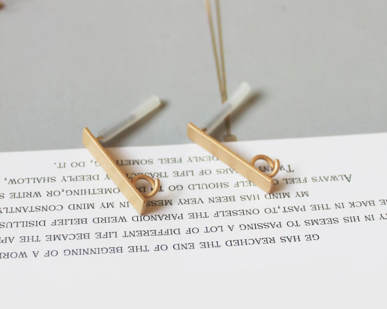 May include: Two gold-colored bar earrings with a small loop at the end. The earrings have a post for insertion into the ear. The background is a light gray surface with text from a book.