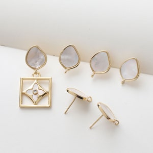 May include: Set of gold-tone earring findings with white shell-like accents. The set includes two pairs of stud earrings and one pair of drop earrings with a square pendant featuring a white flower design.