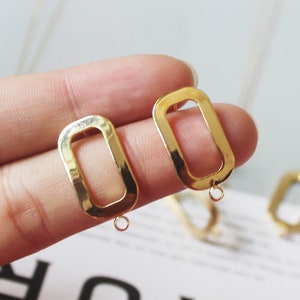 Puede incluir: Un par de pendientes rectangulares dorados con un centro hueco. Los pendientes tienen un pequeño lazo en la parte inferior para sujetarlos a un gancho para la oreja. Los pendientes se muestran sobre un fondo claro.