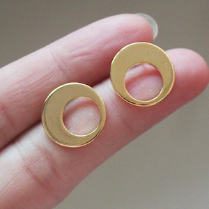 May include: Two gold-colored, circular earrings. Each earring features a solid outer ring with a crescent-shaped cutout, creating a unique, open design. The earrings are shown against a neutral background.