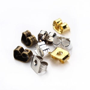 Peut inclure: Assortiment de dos d'oreille en or, argent et bronze. Les dos sont en forme de cœur et ont un petit trou au centre.