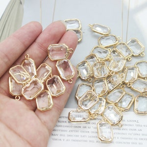 May include: A hand holding a group of clear glass charms with gold-toned metal frames and jump rings. The charms are rectangular in shape with bevelled edges.