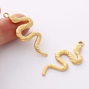 Könnte beinhalten: Zwei goldfarbene Schlangenanhänger mit strukturierten Schuppen und einer Schlaufe zum Befestigen an Schmuck. Die Schlangenanhänger haben eine geschwungene, serpentinenartige Form, vor weißem Hintergrund.
