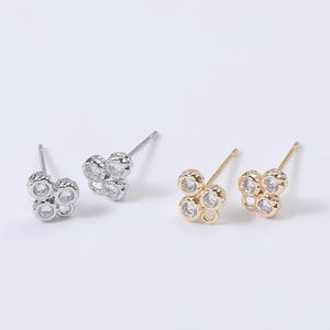 May include: Two pairs of stud earrings. One pair is silver-toned, and the other is gold-toned. Each earring features a cluster of four clear, round gemstones. The earrings are set against a white background.