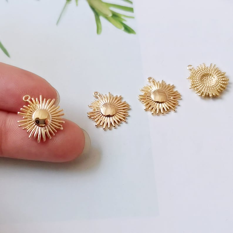 Pu&ograve; includere: Quattro ciondoli a forma di sole con raggio dorati in metallo con un anello in cima. I ciondoli sono disposti in fila su una superficie bianca.