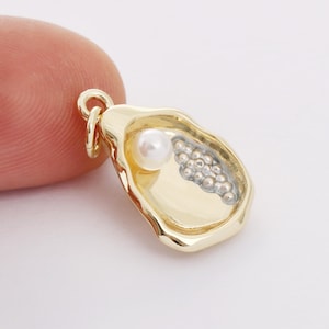 May include: A gold-colored charm in the shape of an oyster shell. The charm features a small white pearl and a cluster of silver-colored beads. The charm has a loop for attaching to a necklace or bracelet.
