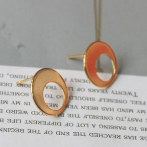 May include: Gold-toned stud earrings with a circular design and a cutout. The earrings have a matte finish with a peach-colored enamel filling the outer ring. The posts are gold-colored.
