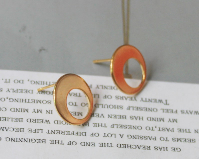 May include: Gold-toned stud earrings with a circular design and a cutout. The earrings have a matte finish with a peach-colored enamel filling the outer ring. The posts are gold-colored.