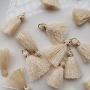 May include: Ivory-colored tassels with gold-colored metal loops and accents. The tassels are made of soft, fringed material and are arranged on a white surface. These decorative tassels are suitable for jewelry making and craft projects.