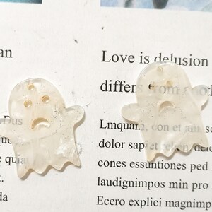Puede incluir: Dos encantos en forma de fantasma blancos con purpurina dorada incrustada en el pl&aacute;stico. Los fantasmas tienen una cara triste con dos ojos y una boca.