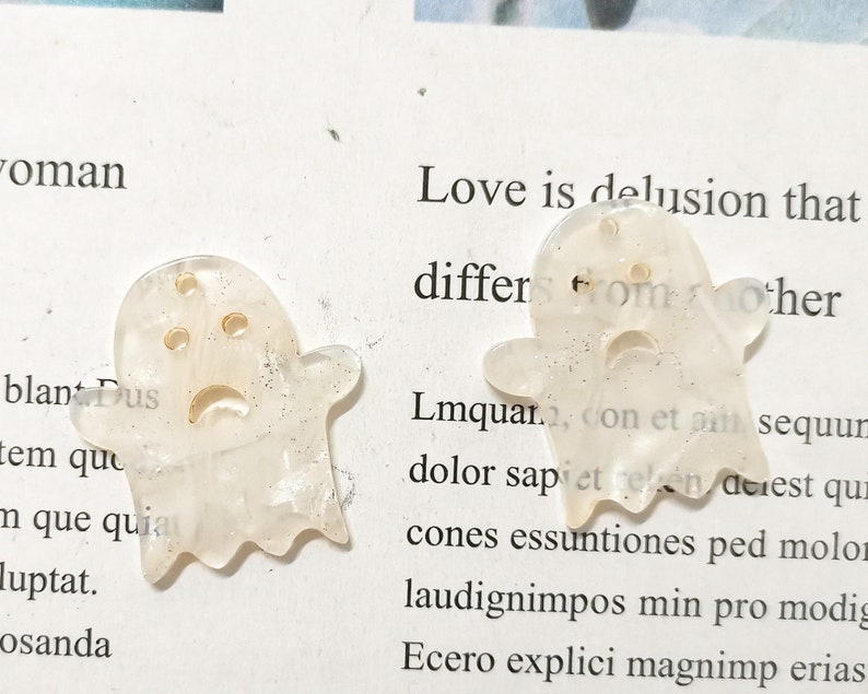 Puede incluir: Dos encantos en forma de fantasma blancos con purpurina dorada incrustada en el pl&aacute;stico. Los fantasmas tienen una cara triste con dos ojos y una boca.