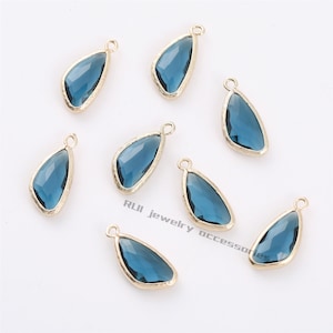Könnte beinhalten: Acht tropfenförmige blaue Glasanhänger mit goldfarbenen Metallrändern. Jeder Anhänger hat oben eine kleine Schlaufe zum Befestigen an einer Kette oder einem anderen Schmuckbestandteil. Die Anhänger sind auf einer weißen Oberfläche angeordnet.