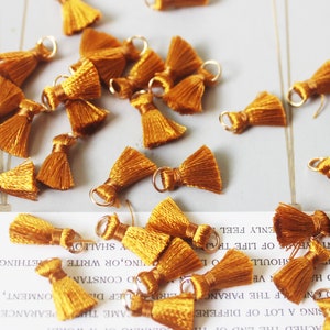 May include: A group of 20 small, mustard yellow tassel charms with gold jump rings. The tassels are made of soft, fluffy yarn and are perfect for adding a touch of whimsy to jewelry, crafts, and home decor.