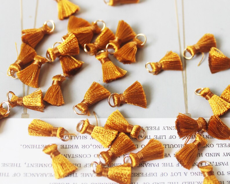 May include: A group of 20 small, mustard yellow tassel charms with gold jump rings. The tassels are made of soft, fluffy yarn and are perfect for adding a touch of whimsy to jewelry, crafts, and home decor.