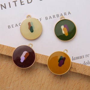 Puede incluir: Cuatro dijes de metal dorado con forma redonda y un bucle para colgar. Cada dije presenta un diseño abstracto diferente con manchas de pintura coloridas en tonos de verde, morado, marrón y amarillo.