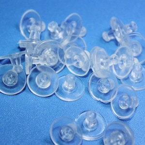 May include: A close-up of a pile of clear plastic earring backs. The backs have a small, round, flat base and a small, round, raised post.