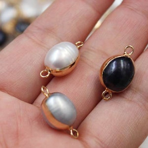 Puede incluir: Primer plano de tres cuentas de perlas ovaladas con detalles metálicos dorados. Una cuenta es blanca, otra gris y otra negra. Cada cuenta tiene un lazo en cada extremo para unir a joyas.