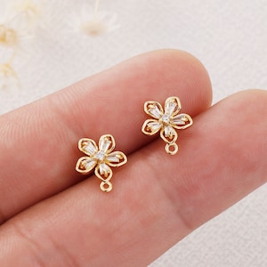 Op de afbeelding: Twee gouden bloemvormige oorbelbedels met heldere zirkonia-stenen. Elke bedel heeft een kleine lus om aan sieraden te bevestigen.